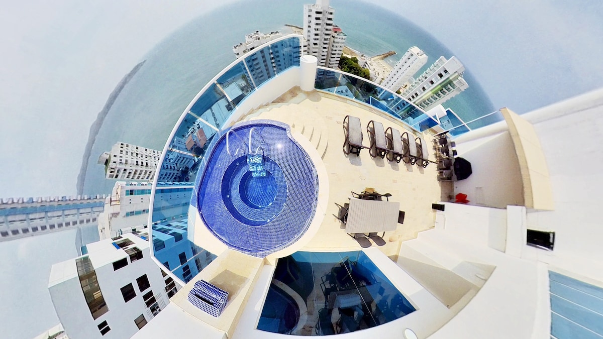 An aerial view captures an inviting terrace featuring a circular swimming pool at its center. Multiple lounge chairs are arranged around the pool, complemented by a dining table set nearby. The expansive ocean and city skyline are visible in the background.