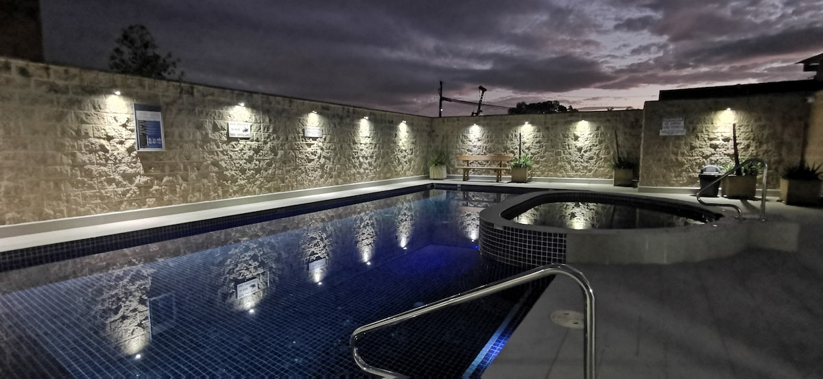 A heated outdoor pool is viewed adjacent to a spa, illuminated by soft lighting on the surrounding stone wall. The calm water reflects the evening sky, creating a serene atmosphere, while potted plants are positioned near the edge, enhancing the tranquil setting.