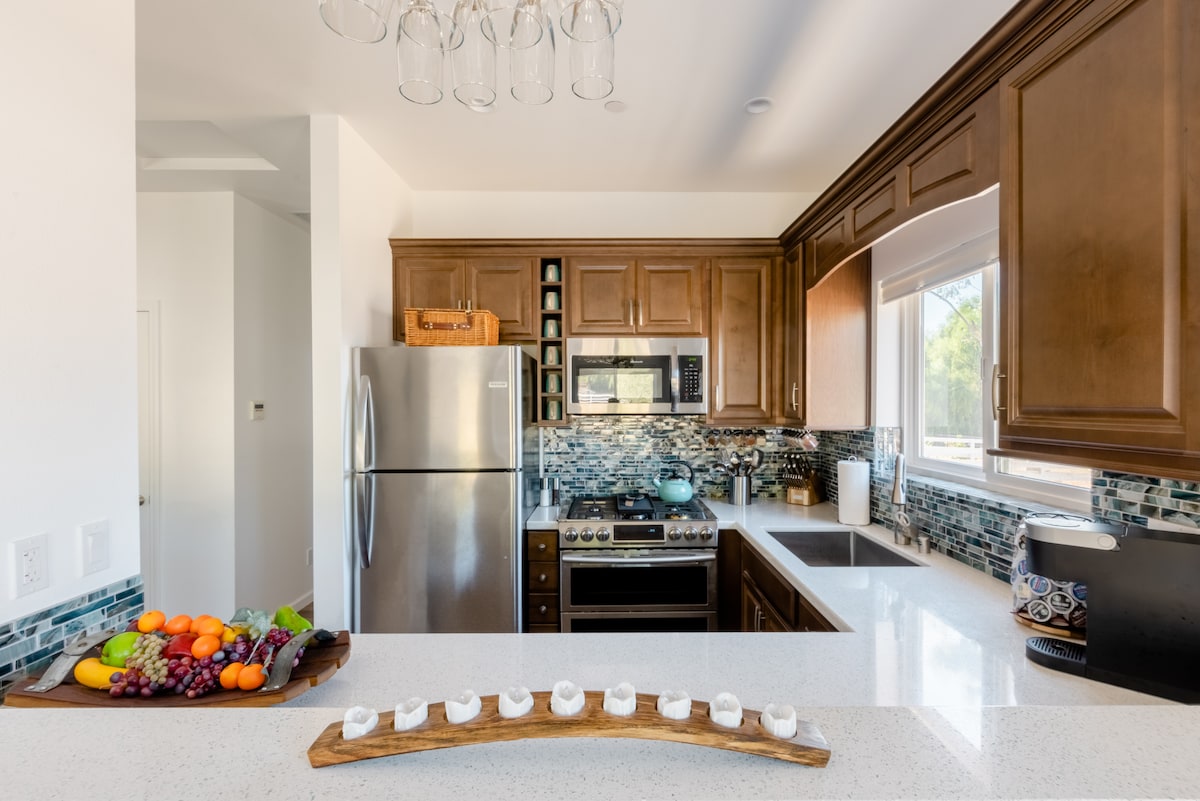 The kitchen features wooden cabinets and a stainless steel refrigerator, complemented by a ceramic stove and sleek countertops. A mixed fruit display is arranged on a wooden tray in the foreground. Natural light comes through the window, enhancing the overall brightness of the space.