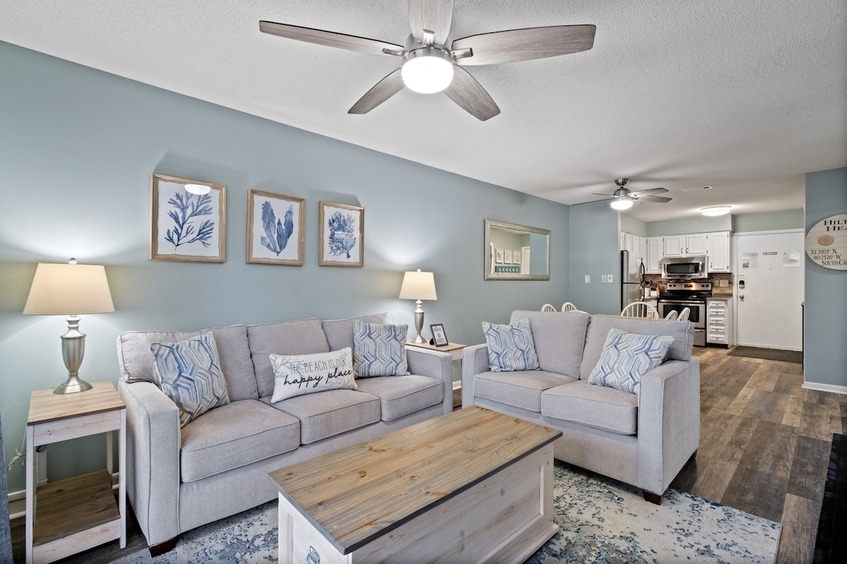 A bright living area features light blue walls and two comfortable sofas, complemented by decorative pillows. A wooden coffee table sits in the center, while lamps provide soft lighting. The open layout leads to a fully equipped kitchen, enhancing the relaxed atmosphere.