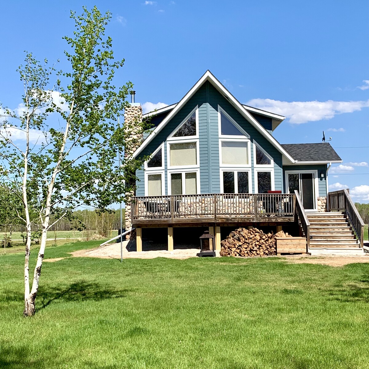 The cottage features a modern design with large windows that provide ample natural light. A spacious deck extends from the structure, overlooking a manicured lawn. A stack of firewood is neatly stored beside the foundation, and a tree adds a touch of greenery to the setting.