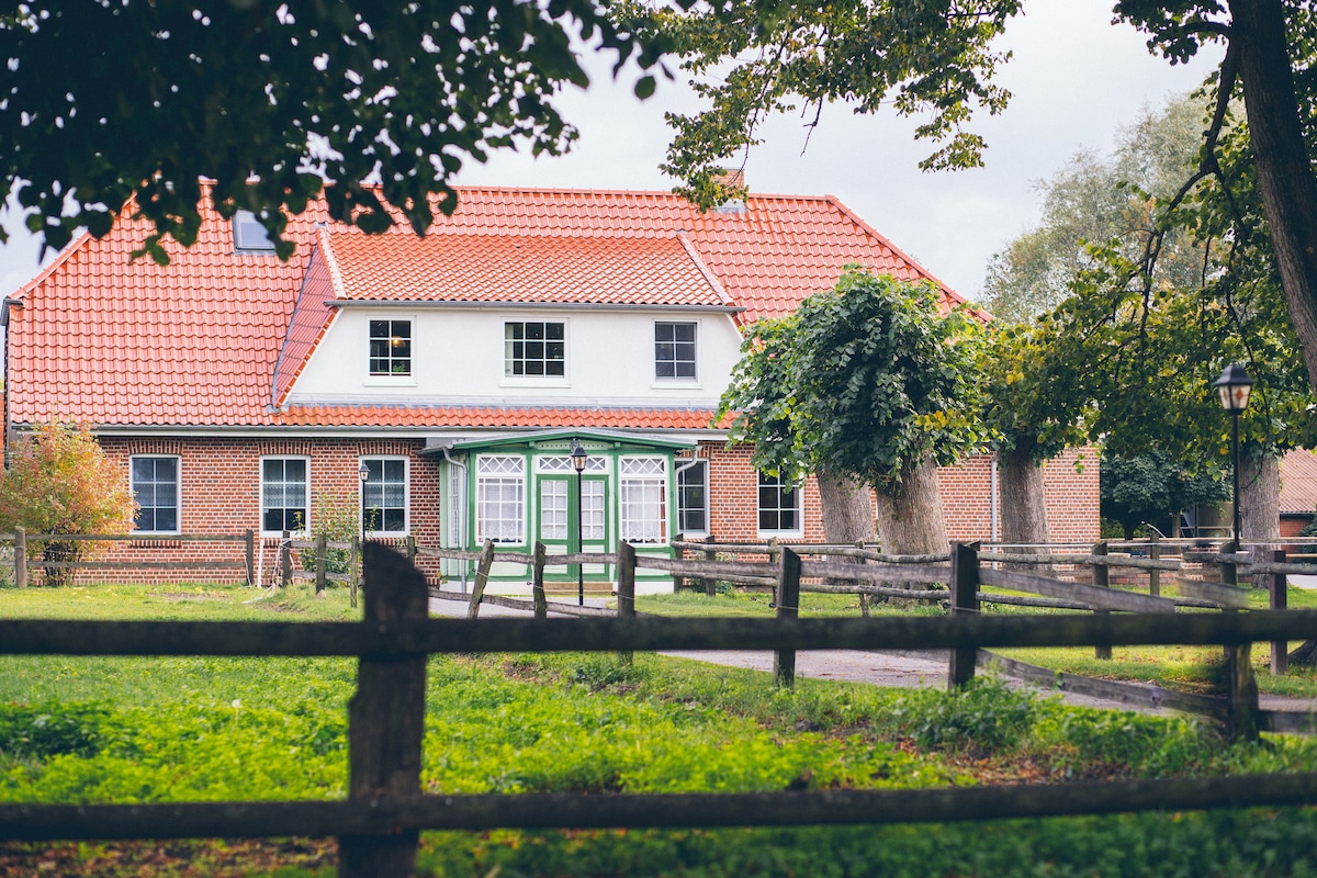 A traditional farmhouse is set within a lush green landscape, featuring a red-tiled roof and decorative white windows. A wooden fence encloses the property, while trees and shrubs provide a natural backdrop, contributing to the serene atmosphere of the rural setting.