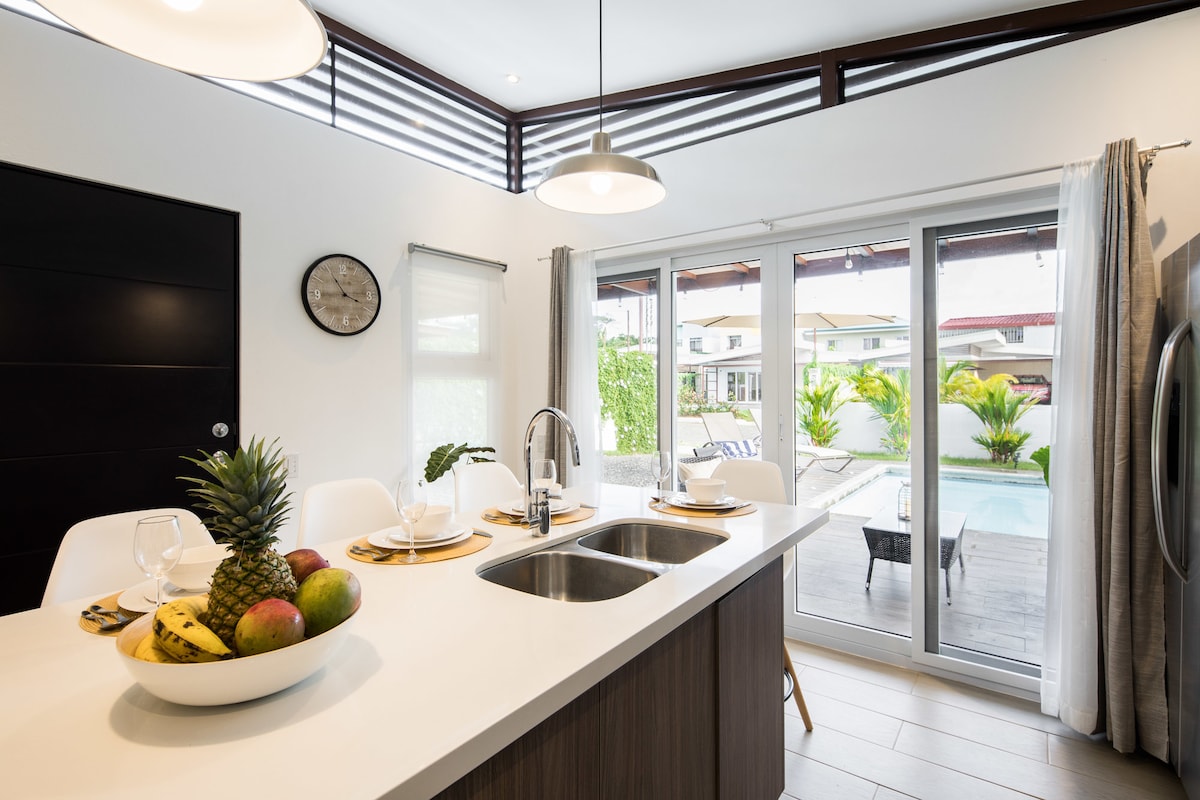 A modern kitchen features a central island with a sink, surrounded by white seating. Fresh fruit, including bananas and pineapple, is displayed on the island. Large glass doors open to a private pool area, allowing for ample natural light.