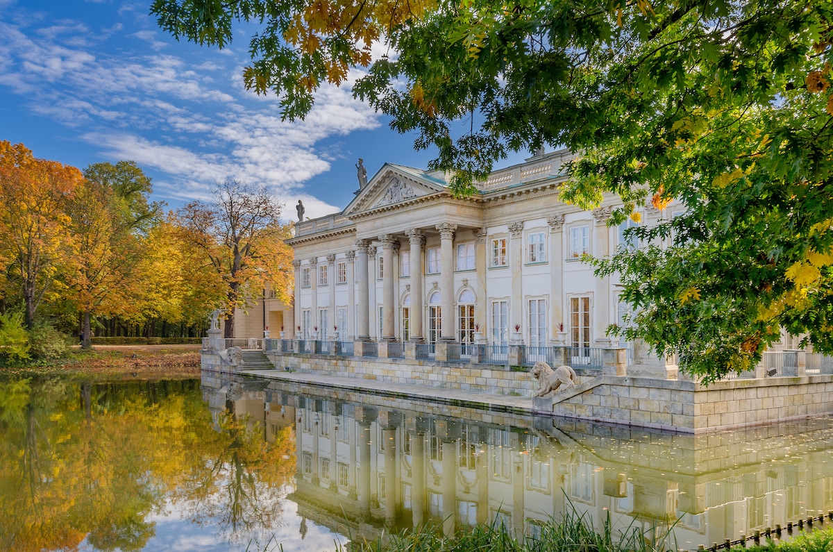 A historic building is reflected in a serene body of water, surrounded by lush greenery and trees displaying autumn colors. The architecture features large columns and detailed sculptures, enhancing the elegant appearance of the structure. Clear blue skies complement the tranquil scene.