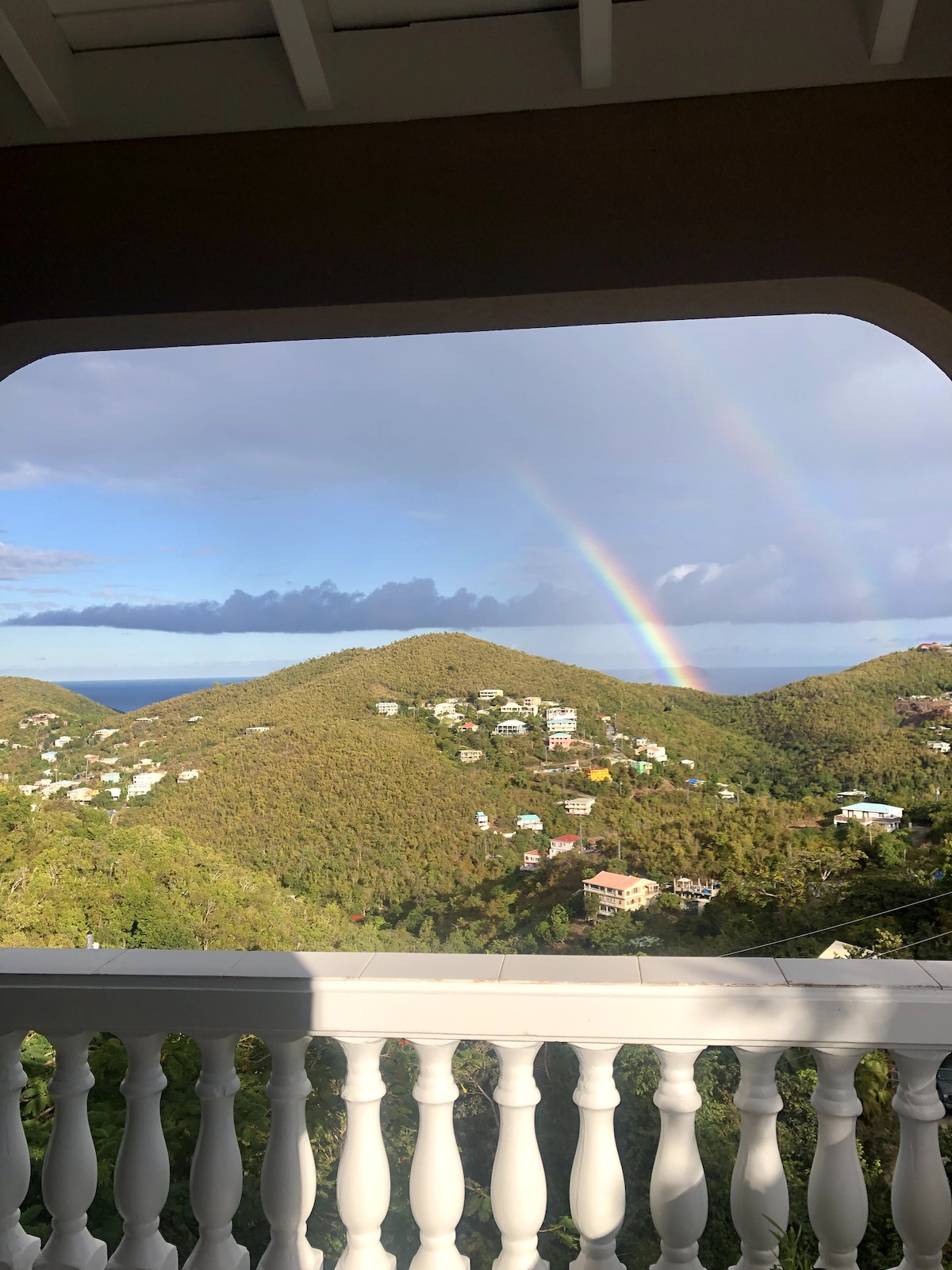 A covered balcony offers a stunning view of rolling green hills and the Caribbean Sea. A vibrant rainbow arches over the landscape, accentuating the scenic beauty of the surrounding natural environment. The elegant balustrade frames the picturesque vista.