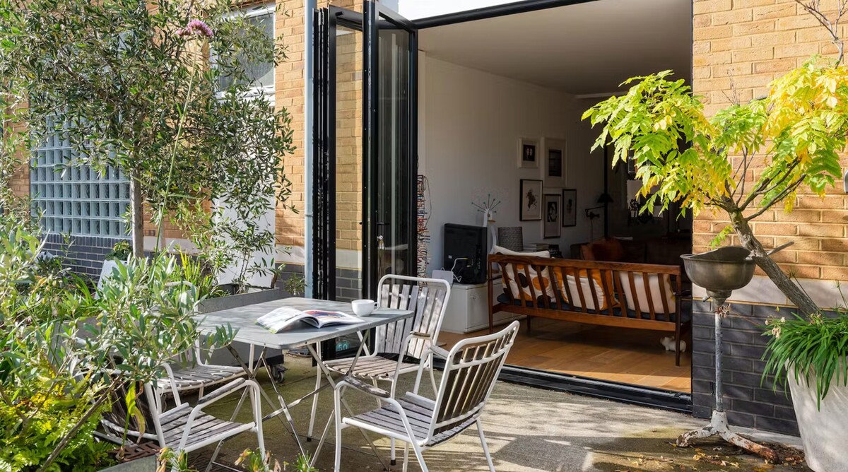 An outdoor terrace is presented with a small round table and four metal chairs, surrounded by greenery. The open glass doors reveal a tastefully furnished interior with a comfortable seating area. Natural light enhances the inviting ambiance of the connected spaces.
