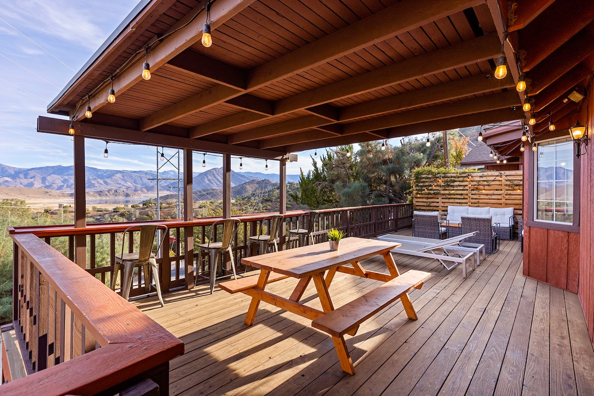 An outdoor patio area features a wooden picnic table and multiple seating arrangements under string lights. The space offers expansive views of distant mountains and the surrounding landscape, inviting outdoor gatherings and relaxation.