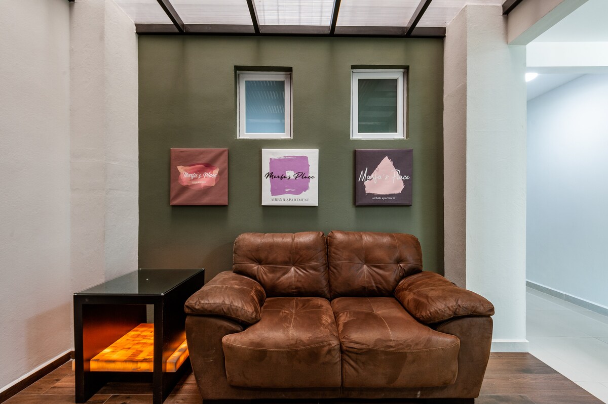 A spacious seating area features a brown leather sofa positioned against a green wall. Abstract art pieces adorn the wall above, complementing the modern design. Natural light filters in through the windows, adding warmth to the space.