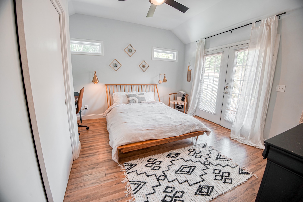 A bedroom features a wooden bed with white linens and a decorative rug. Two wall sconces provide soft lighting beside the bed. Natural light enters through double glass doors, which lead to the outside. Simple decor and wooden flooring create a serene environment.