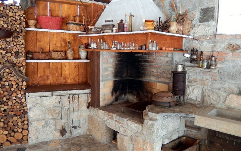 A rustic kitchen area features a stone fireplace with a traditional cooking surface. Shelves display various glass jars and cookware, complemented by woven baskets. A stack of firewood is arranged against the stone wall, enhancing the room's warm, earthy character.