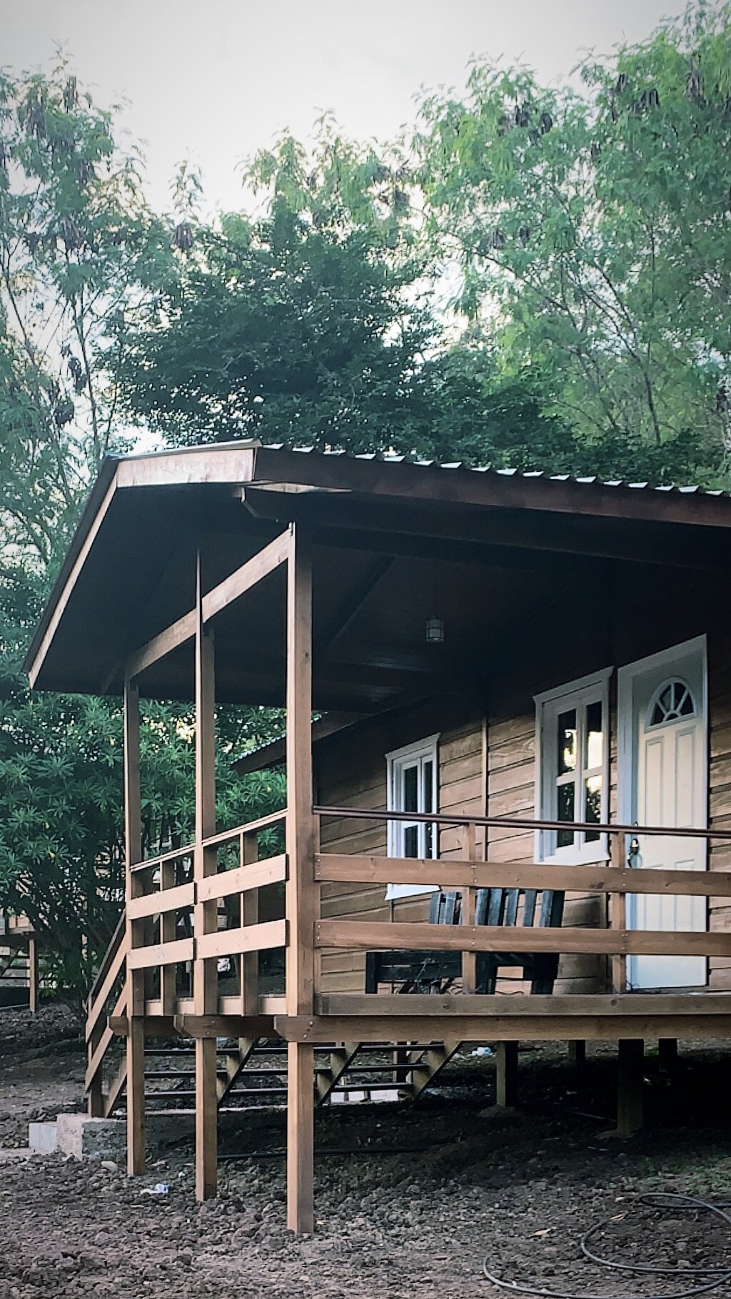 A wooden cabin with a covered porch is surrounded by greenery. The porch features steps leading down and showcases two chairs positioned for outdoor seating. Large windows allow natural light, and the building's simple design harmonizes with the natural landscape.