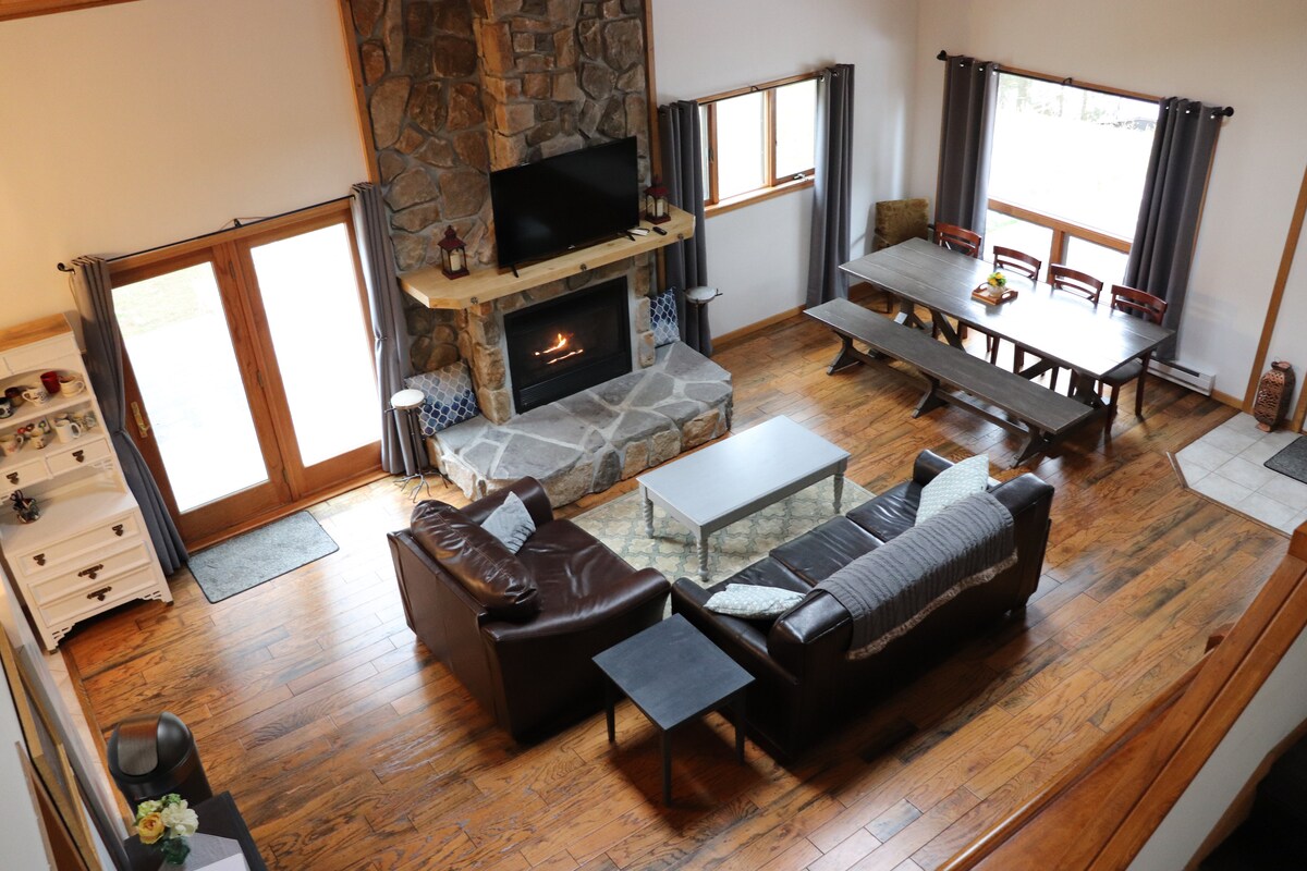 The open living space features a stone fireplace, surrounded by comfortable seating arrangements. Large windows allow natural light to fill the area, which is furnished with a dining table and additional seating. A coffee table sits centrally, enhancing the room's functionality.