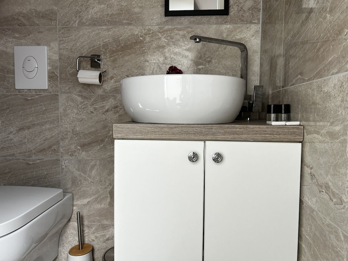 A modern bathroom features a white, round basin atop a light wooden countertop. Small toiletries and a rectangular mirror are situated beside it. A white toilet is visible to the left, enhancing the clean and contemporary aesthetic of the space.