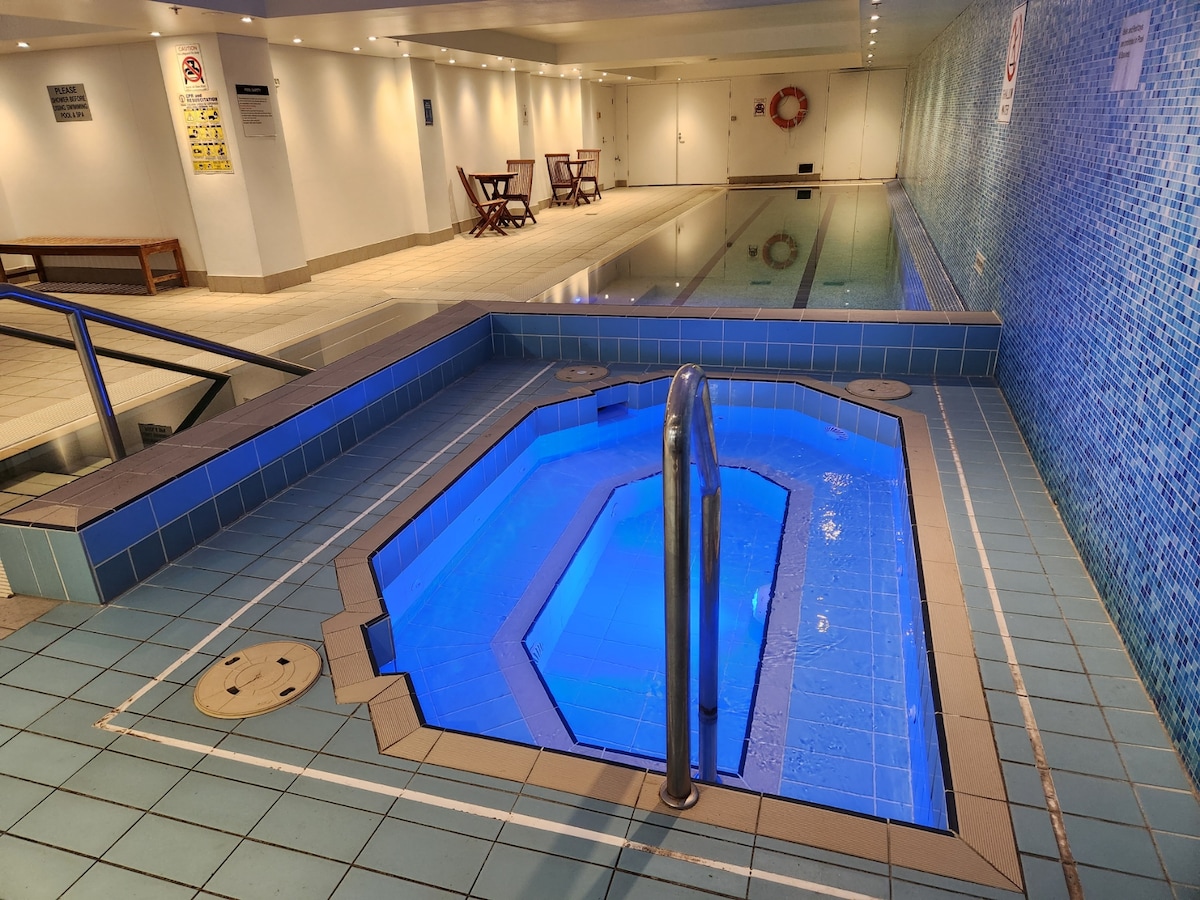An indoor heated lap pool is presented alongside a jacuzzi featuring blue lighting. The tiled surface surrounds both the pool and jacuzzi, with seating available in the background. The facility is well-lit, highlighting the clean lines and relaxing ambiance.