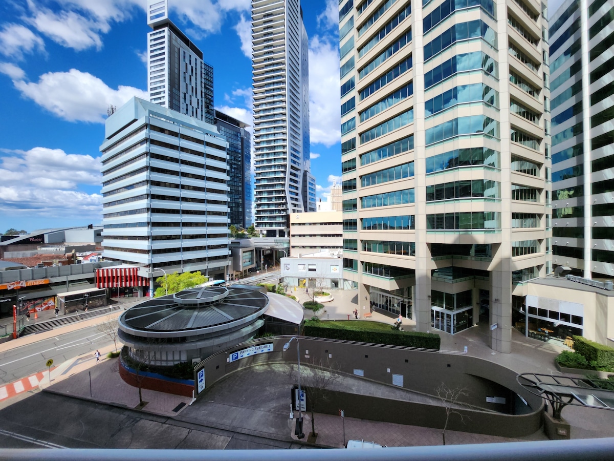 A view from a balcony overlooks a bustling urban landscape. Tall buildings of varying designs are visible, showcasing modern architecture. A circular structure is situated below, with a road running alongside. Clear skies with scattered clouds create a bright atmosphere.