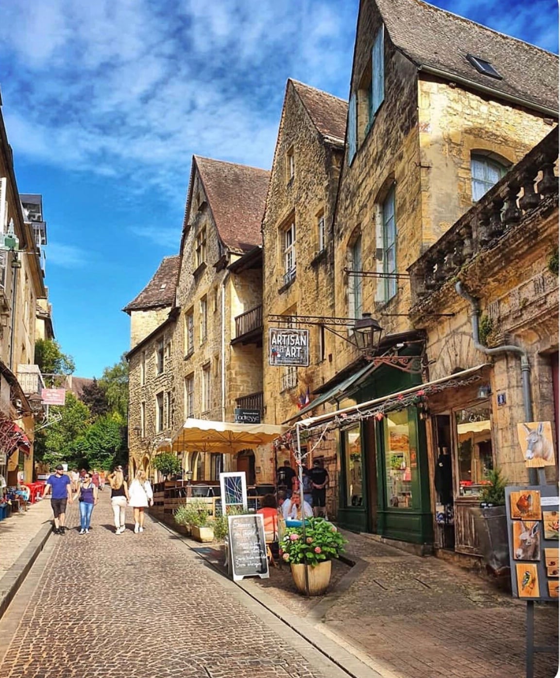 A charming street lined with stone buildings showcases a blend of artistic shops and cafés. People stroll along the cobblestone pathway under a clear blue sky, while greenery and outdoor seating add to the inviting atmosphere of this historic area.