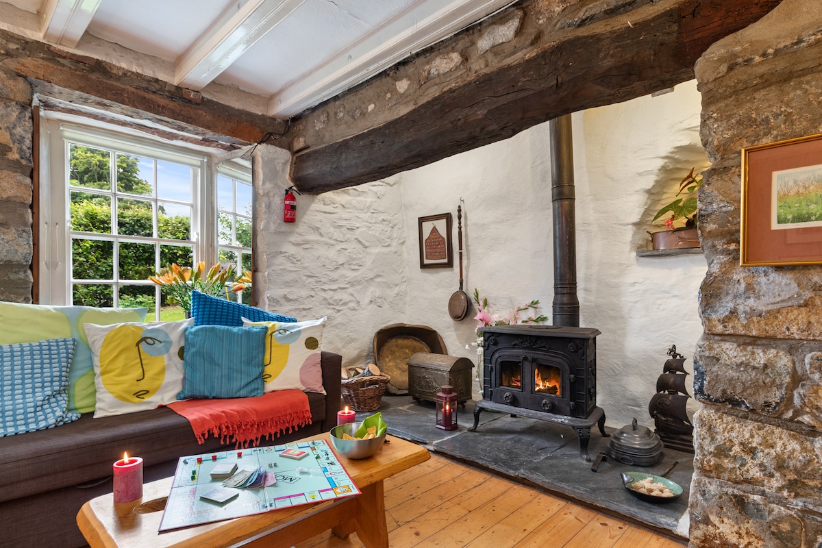 A cozy living space is featured with a rustic wooden beam overhead and a stone wall. A wood-burning stove is positioned against one wall, providing warmth. A comfortable sofa adorned with colorful cushions faces a small wooden table, set with a board game and candles.