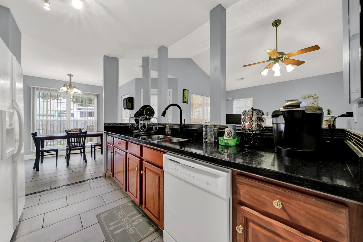 A modern kitchen is presented with dark countertops and wooden cabinetry. Bright lighting illuminates the space, highlighting a dining area with a small table and chairs. A large refrigerator and dishwasher are visible, alongside a coffee maker and organized storage for kitchen essentials.