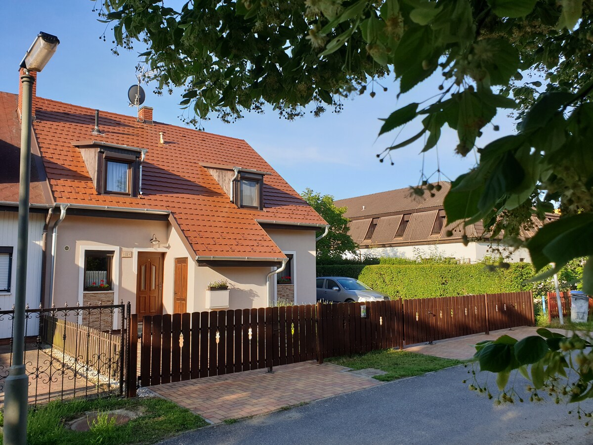 A charming house is framed by green foliage, showcasing a red-tiled roof and a wooden fence. A pathway leads to the main entrance, where a door provides access. The surrounding area is lined with greenery, with a vehicle parked nearby.