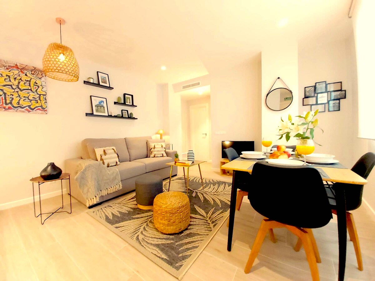 A modern living area features a soft gray sofa adorned with decorative cushions. A wooden table is set for dining, showcasing plates and glasses. Wall art and family pictures create a personalized touch. Natural light fills the room through a large window.