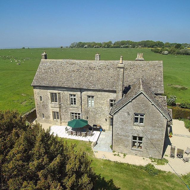 An aerial view of King John's Palace showcases the historic stone structure surrounded by lush green fields. A large patio area is visible, featuring outdoor seating and an umbrella. The property is accompanied by mature trees, enhancing the serene rural setting.