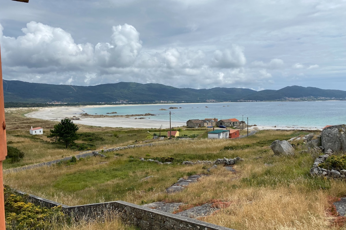 A panoramic view showcases a serene beach and coastline, with gently rolling waves meeting the sandy shore. The tranquil landscape includes a patch of green grass in the foreground, leading to colorful buildings dotting the shore against a backdrop of rolling hills and scattered clouds.