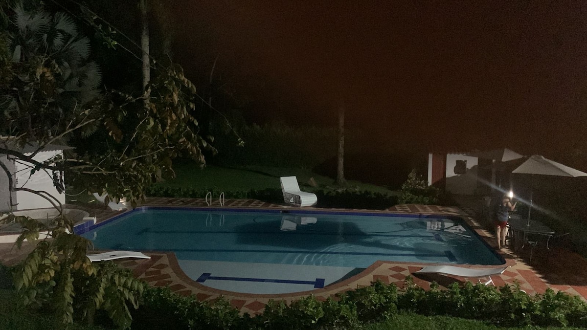 A tranquil outdoor pool area is visible, surrounded by lush greenery. Soft lighting reflects gently on the water's surface, and a shaded seating area is positioned nearby. Two lounge chairs are arranged by the pool, enhancing a serene atmosphere conducive to relaxation.