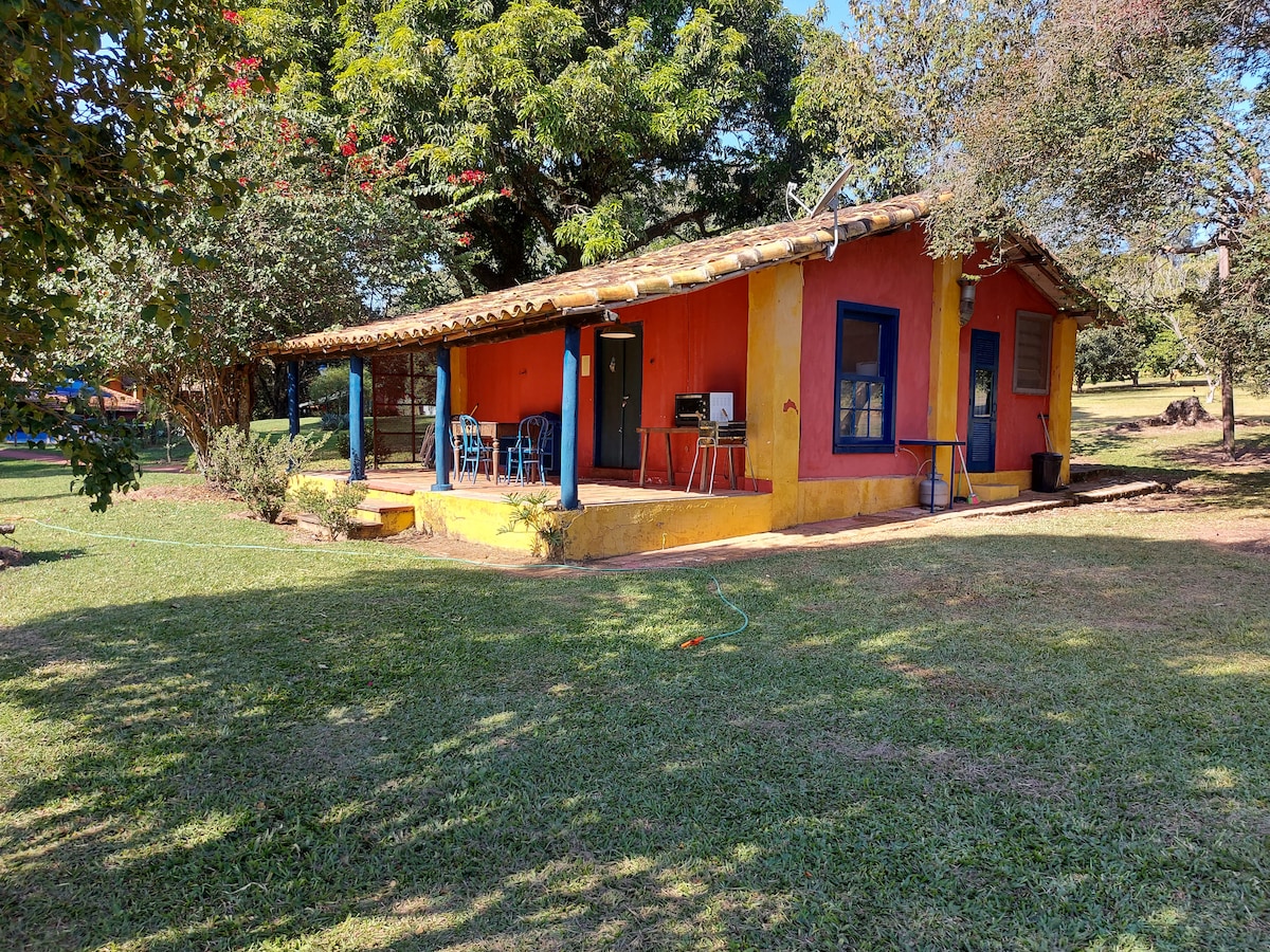 The exterior of a colorful cottage is featured, surrounded by lush greenery. The structure has a yellow and orange facade with a traditional tiled roof. A small porch is visible, furnished with blue chairs and a table, providing a welcoming space to enjoy the outdoors.