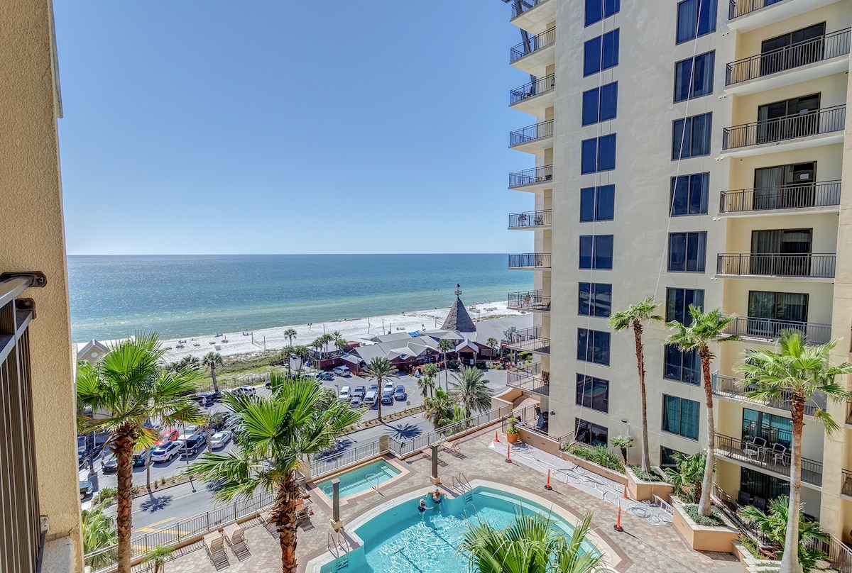 The view features the expansive Gulf of Mexico with its vibrant blue waters, alongside a community pool surrounded by palm trees. Below, a beach access path leads to the sandy shore. A taller building stands adjacent to the pool area, enhancing the coastal setting.