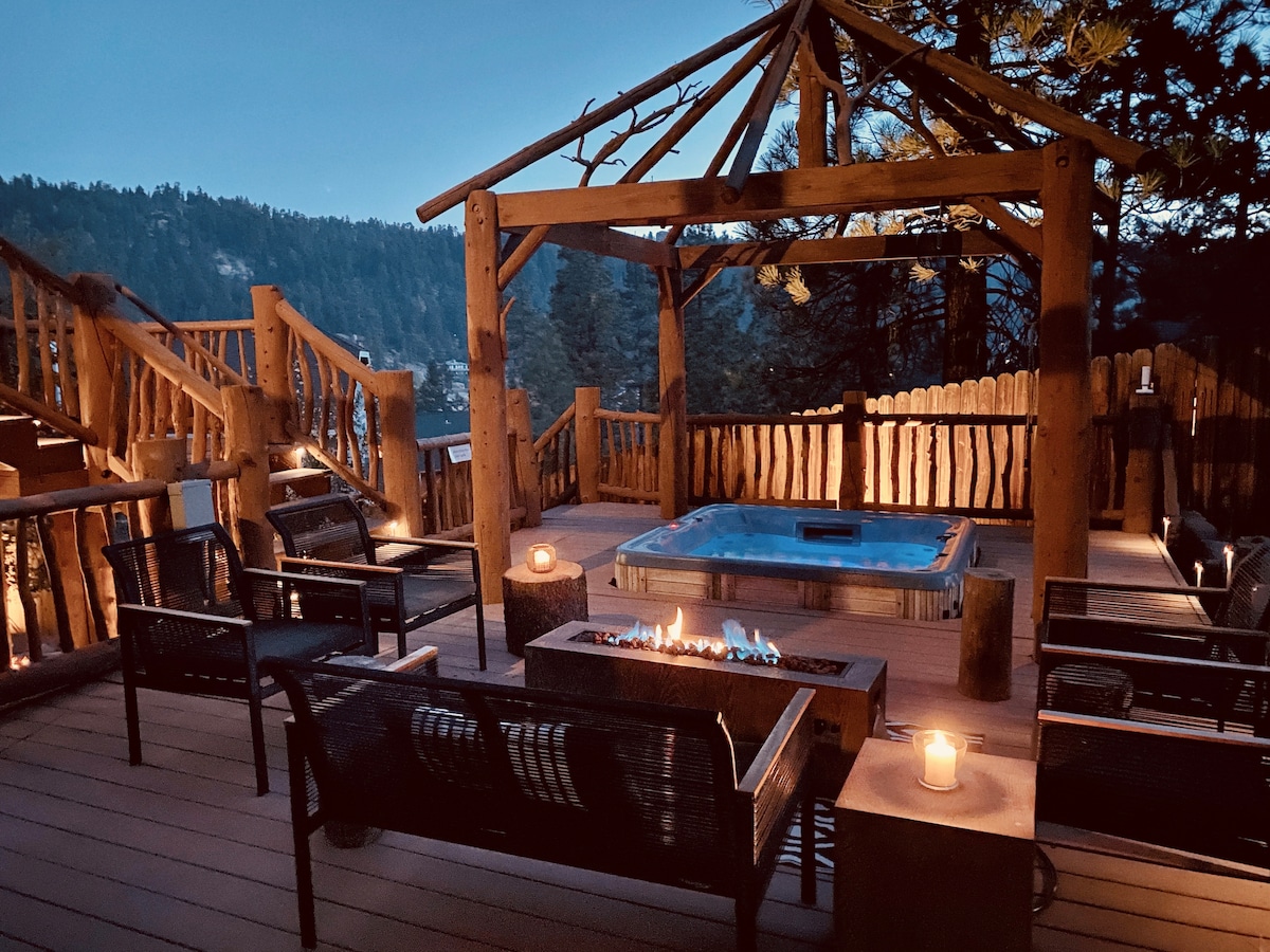 An outdoor spa area features a hot tub surrounded by rustic wooden posts, with a fire pit at the center. Soft lighting from candles creates a warm ambiance, while seating is provided on woven lounge chairs. Scenic mountain views are visible in the background.