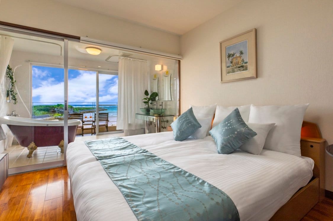 A bright bedroom features a spacious bed adorned with decorative pillows. Large glass doors provide ocean views and natural light, leading to a balcony. A cozy seating area is visible, complemented by a bathtub positioned near the window for relaxation.