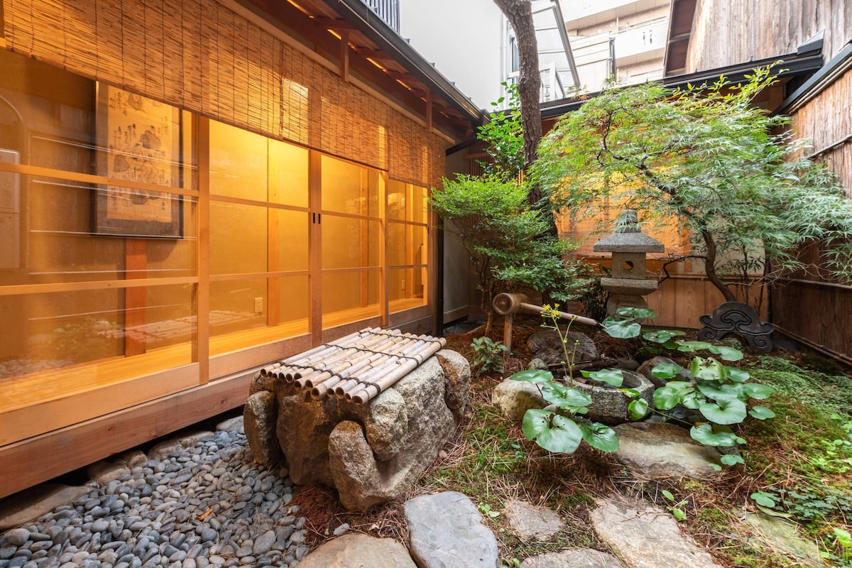 A serene courtyard features a traditional Japanese garden design, with lush greenery, moss, and arranged rocks. A stone lantern stands beside a pond adorned with lily pads. Sunlight filters through bamboo shades, creating a warm and inviting environment that emphasizes tranquil outdoor living.