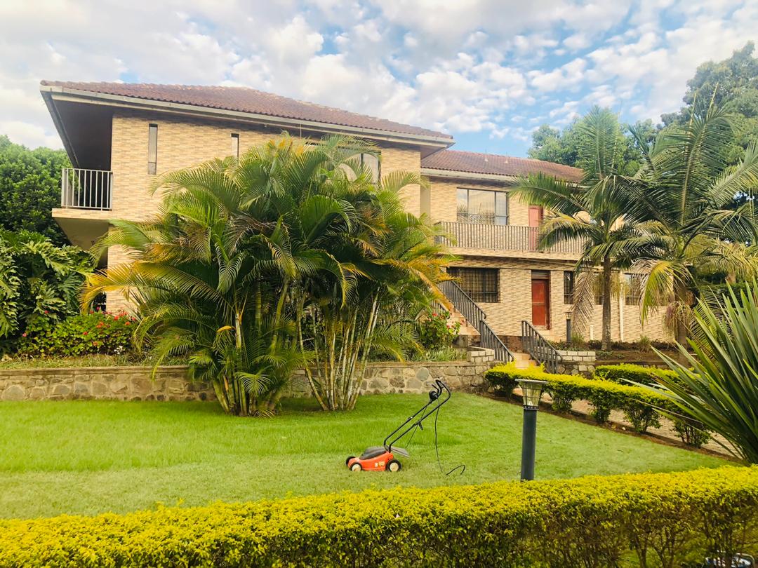 A two-story brick building is surrounded by a well-maintained garden featuring lush greenery and palm trees. A manicured lawn is visible, with a lawn mower placed nearby. Pathways leading to the entrances are bordered by vibrant yellow flowers and shrubs.