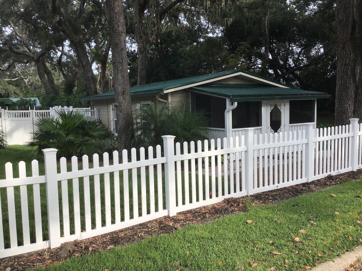 A charming cottage is surrounded by a white picket fence and features a green roof. Lush greenery, including palm trees and moss-draped oaks, is visible in the yard, contributing to a secluded feel. The entrance is marked by a light-colored door with a decorative window.