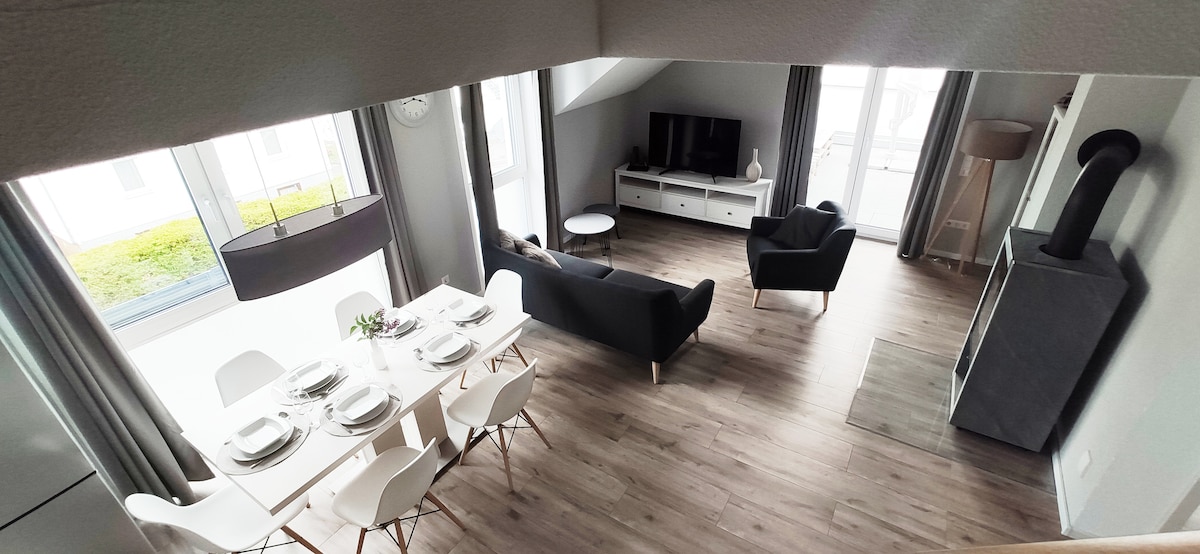 A spacious living area is depicted, featuring a dining table set for six, complemented by modern white chairs. A comfortable couch and two armchairs are arranged around a sleek television unit. Natural light floods the room through large windows, highlighting the cozy ambiance.