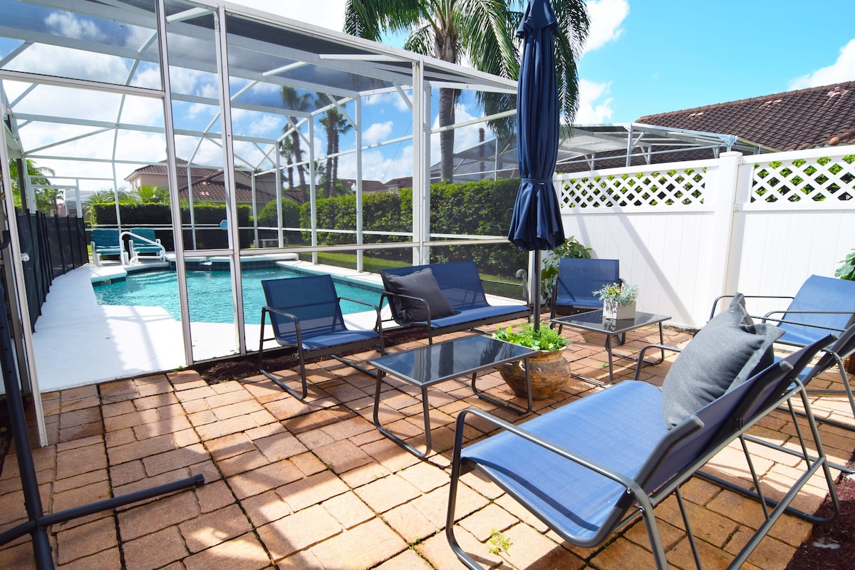 An outdoor pool deck is surrounded by a screened enclosure, featuring several lounge chairs and a patio table. A large umbrella provides shade, while potted plants add a touch of greenery. The private pool is visible in the background, creating a relaxed outdoor setting.