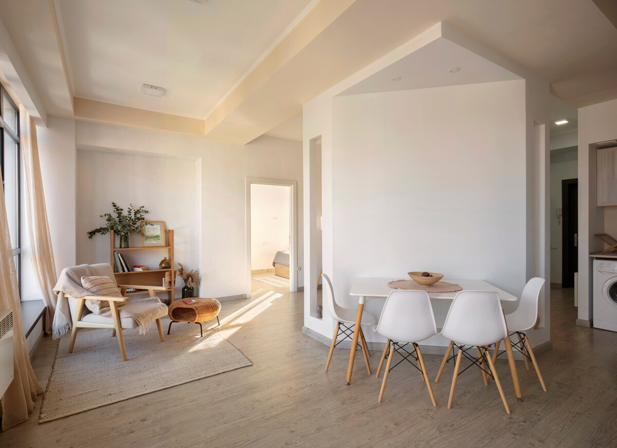 A modern living and dining area features light wood flooring and walls painted in neutral tones. A dining table surrounded by chairs is visible, along with a cozy seating area including a chair and a woven basket. Natural light illuminates the space through large windows.