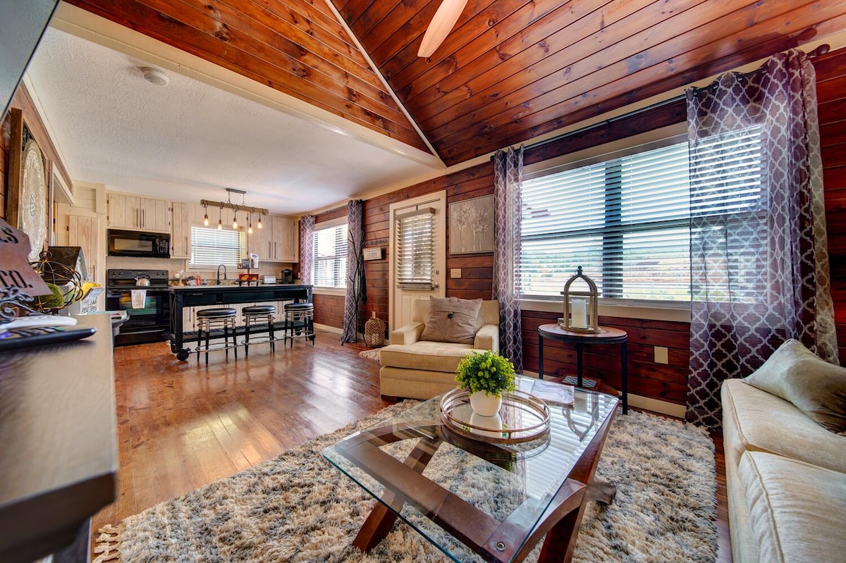 An open living area features wooden ceilings and a cozy seating arrangement, including a light sofa and armchair. A glass coffee table sits on a plush area rug, while a dining table is visible in the kitchen area alongside modern appliances and ample windows providing natural light.