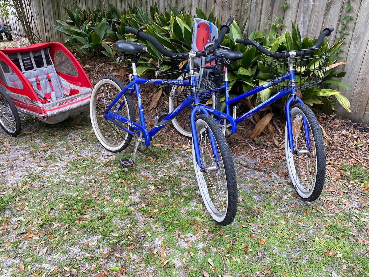 Two blue bicycles are parked next to a red bike trailer, surrounded by lush greenery and a wooden fence. Both bicycles feature front baskets and have sturdy frames, offering a convenient option for exploring the nearby beach and neighborhood.