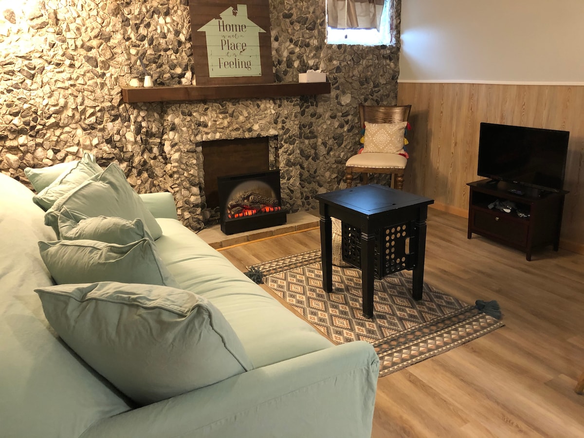 A cozy living area is highlighted by a stone accent wall and an electric fireplace beneath a wooden mantel. A soft blue sofa with numerous cushions faces a small coffee table, positioned on a patterned rug. A television is located beside a wooden cabinet.