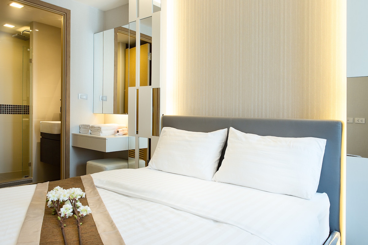 A spacious bedroom features a queen size bed with crisp, white linens and two decorative pillows. The sleek headboard complements the contemporary design. A light-colored accent wall creates a soothing ambiance, while a connected bathroom is partially visible, showcasing modern amenities.