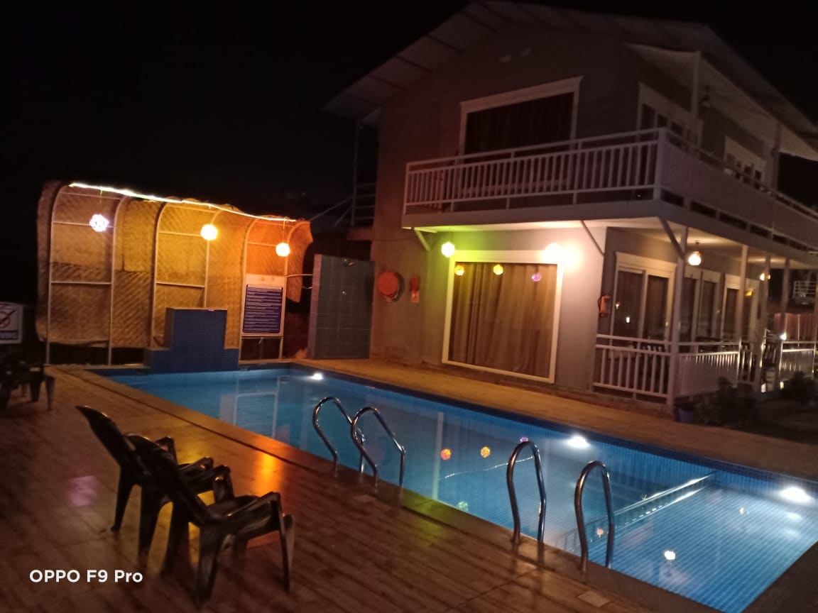 An outdoor swimming pool is illuminated at night, surrounded by a wooden deck and lounge chairs. A cozy thatched hut is visible nearby, adorned with colorful lights. The structure behind features large windows reflecting the soft glow from the pool area.