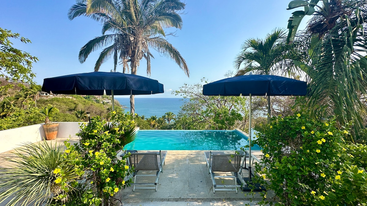 An inviting infinity pool, framed by lush greenery, offers stunning ocean views. Two lounge chairs are positioned under large umbrellas, providing shade. Vibrant yellow flowers add a touch of color to the serene outdoor space, creating a relaxing environment.