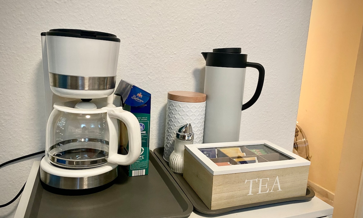 A coffee maker with a glass carafe sits on a tray next to a tea box labeled 'TEA'. A decorative thermos and a cylindrical container with a lid are also present. A milk carton is placed nearby, and a small salt shaker completes the arrangement.