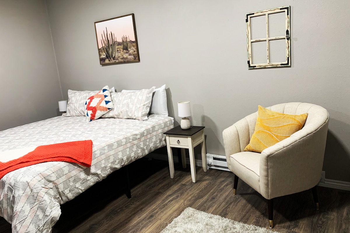 A spacious sleeping area features a king-size bed adorned with patterned bedding and decorative pillows. A comfortable armchair sits nearby, accompanied by a small nightstand with a lamp. A framed photograph and a window frame artwork add visual interest to the neutral-colored walls.