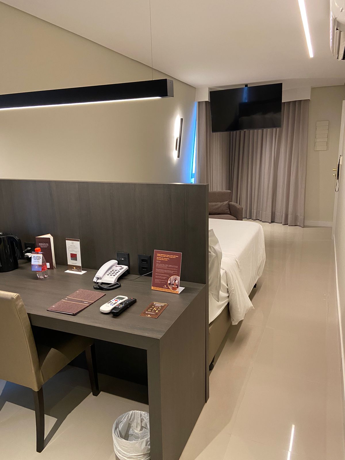 An inviting executive suite features a sleek desk with a chair, positioned opposite a spacious bed. A flat-screen television is mounted on the wall, and soft curtain panels frame a window, allowing natural light to filter in. Neutral tones contribute to a calm ambiance.