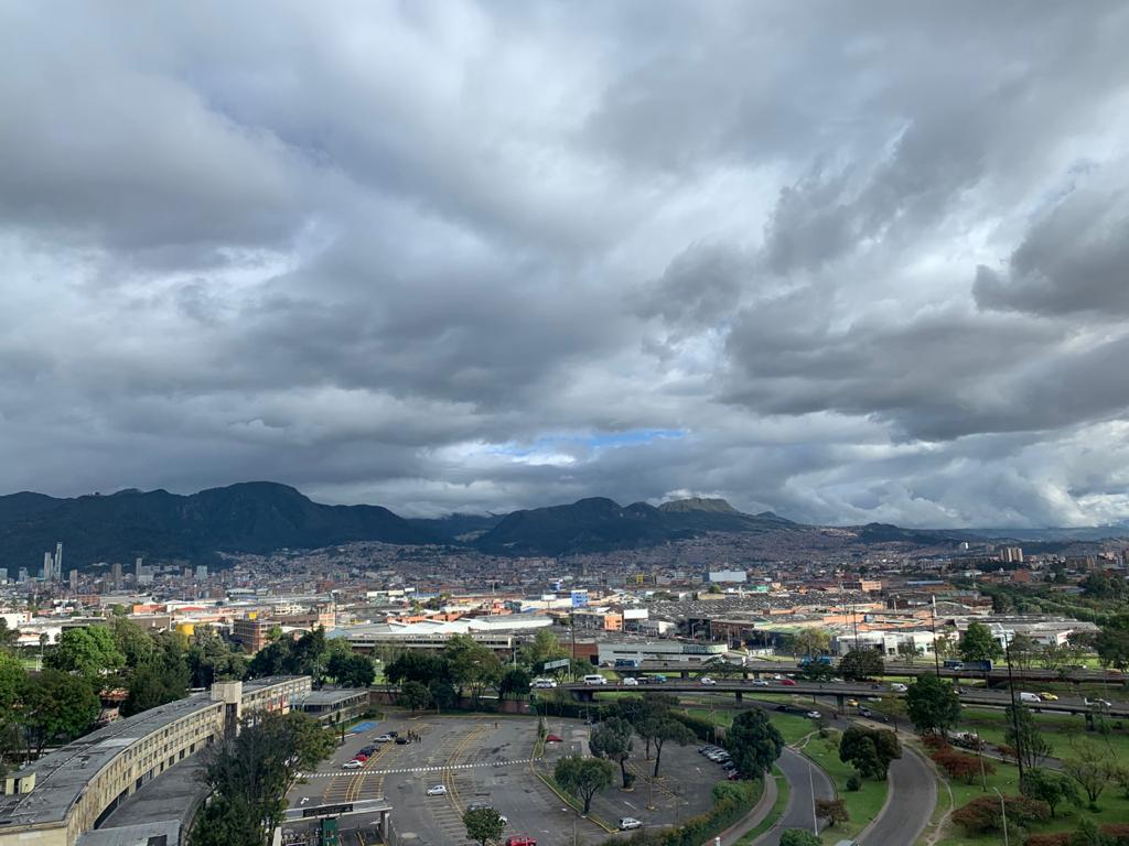 A panoramic view captures the sprawling cityscape of Bogotá, framed by dramatic mountains under a cloudy sky. The scene includes green parks and urban structures, illustrating the vibrant blend of nature and city life.