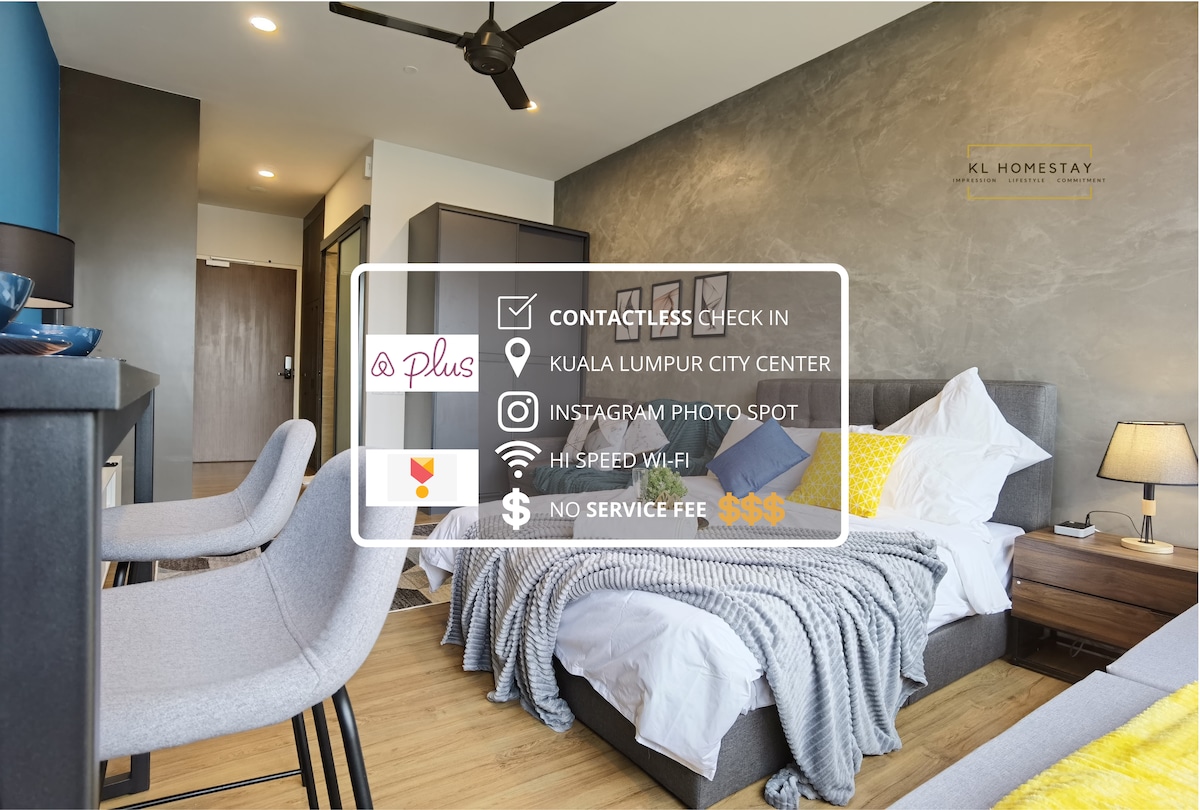 A modern studio bedroom is featured, showcasing a queen-size bed with a light duvet and pillows. Two gray chairs are positioned nearby, and wooden flooring adds warmth to the space. Natural light enters through the window, creating a bright and inviting atmosphere.