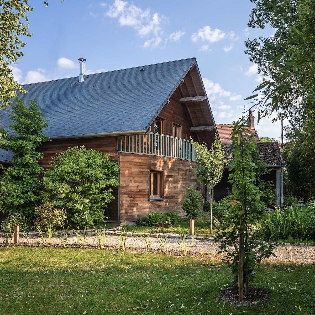 A rustic wooden house is framed by lush greenery and garden pathways. A charming balcony is positioned above the entrance, with trees and bushes adding a natural touch. The sloped roof and warm wood tones contribute to a welcoming presence within the surrounding landscape.