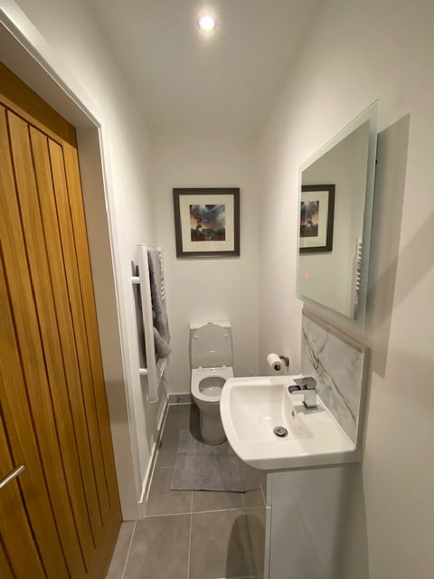 A modern ensuite shower room is presented, featuring a white toilet and a sleek sink with a chrome faucet. Neutral wall tiles complement the space, which is illuminated by recessed lighting. A large mirror and two framed artworks add a touch of design, while a towel rack is mounted on the wall.